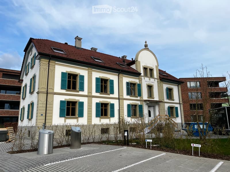 A two-story residential building with a red-tiled roof, white and beige facade, green shutters, and multiple windows. The building features a small dome-shaped turret and is surrounded by a paved area with trash bins and a sign indicating private property