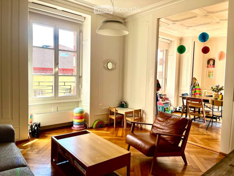 A modern, well-lit living room with wooden flooring, a large window offering a view of the roof, a leather armchair, a sofa, a coffee table, a dining area with a table and chairs, and colorful decorative items.