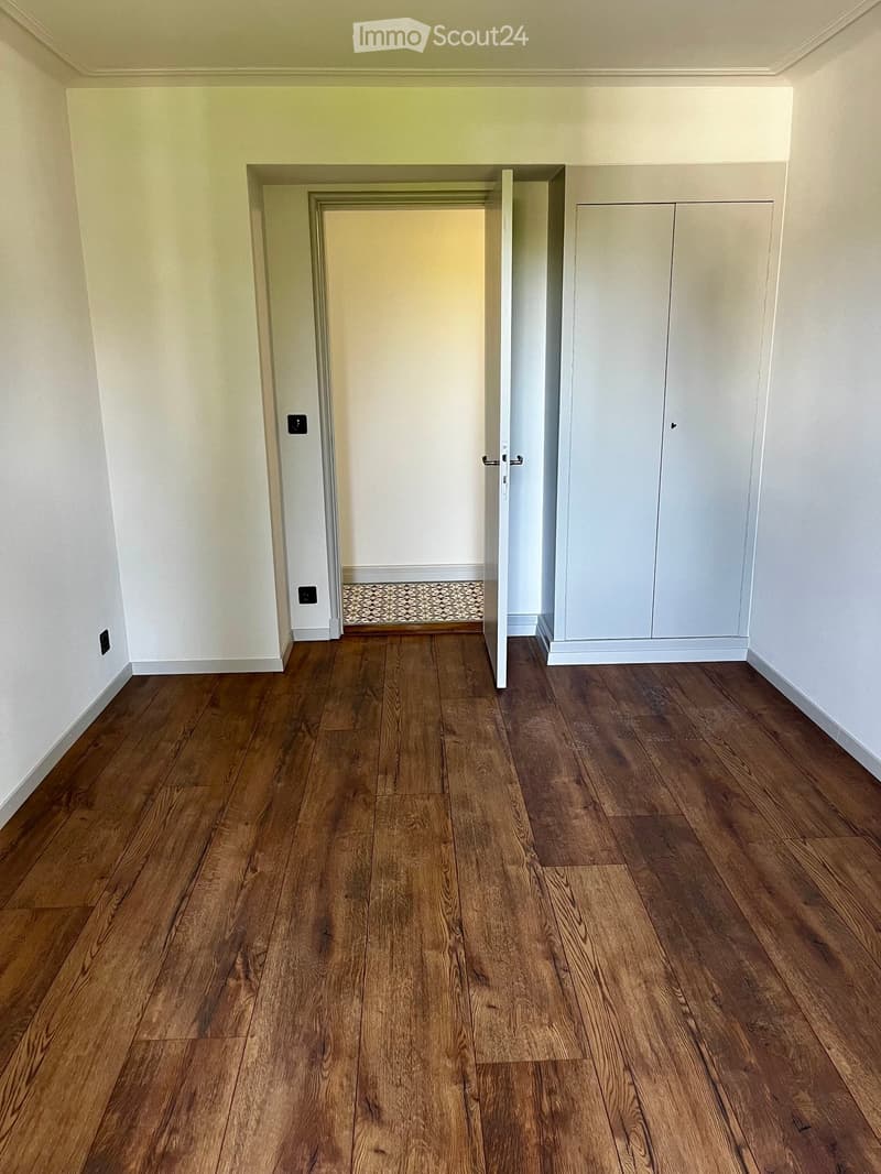 Empty room with wooden flooring, white walls, and a built-in closet with double doors. A small patterned rug is visible in the adjacent room through an open doorway.