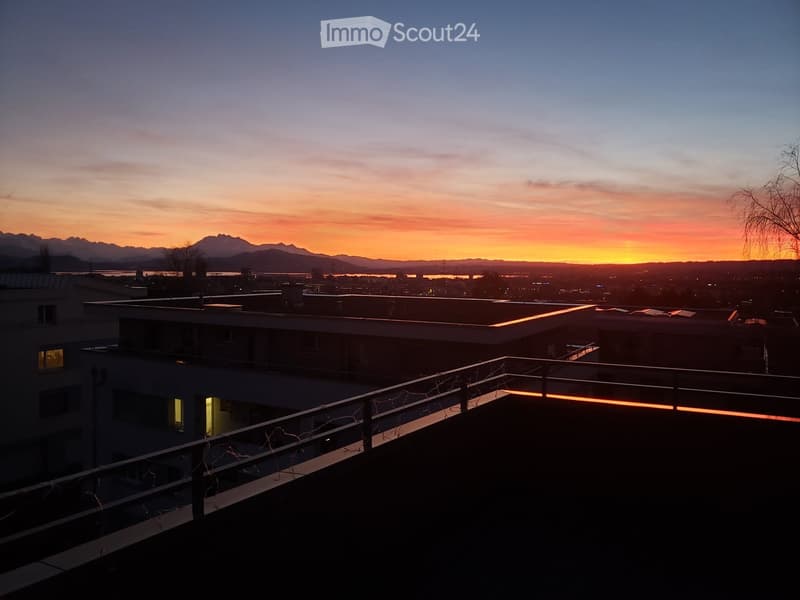 Balcony with a railing, overlooking a cityscape at sunset with mountains in the background.