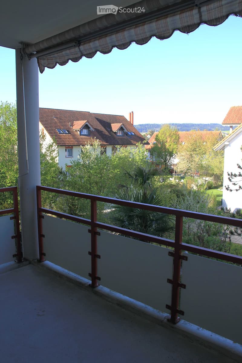 Balcony with a view of a residential area, featuring a wooden railing, a striped awning, and a clear blue sky.