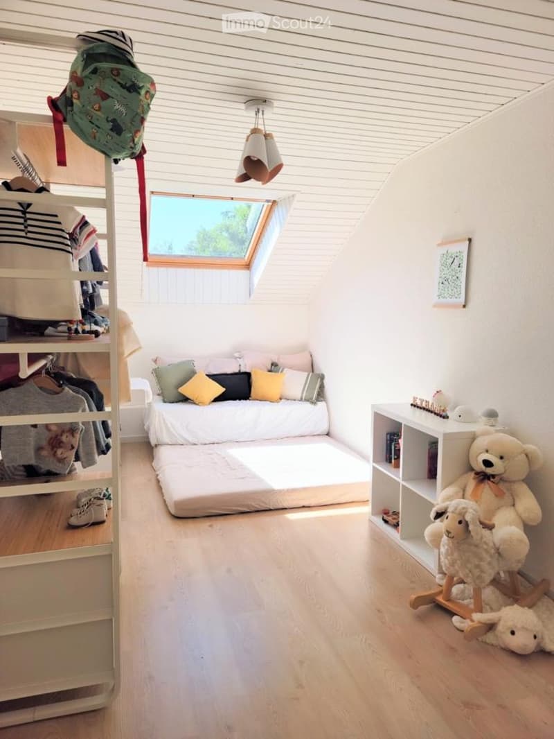 Cozy attic bedroom with a lofted bed, pillows, a small shelf, a rocking horse, and a teddy bear. The room features wooden flooring, white walls, and a skylight window.