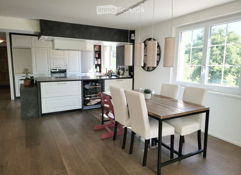 Modern kitchen with white cabinets, dark countertops, and a central island. A wooden dining table with white chairs is placed near large windows, with pendant lights hanging above.