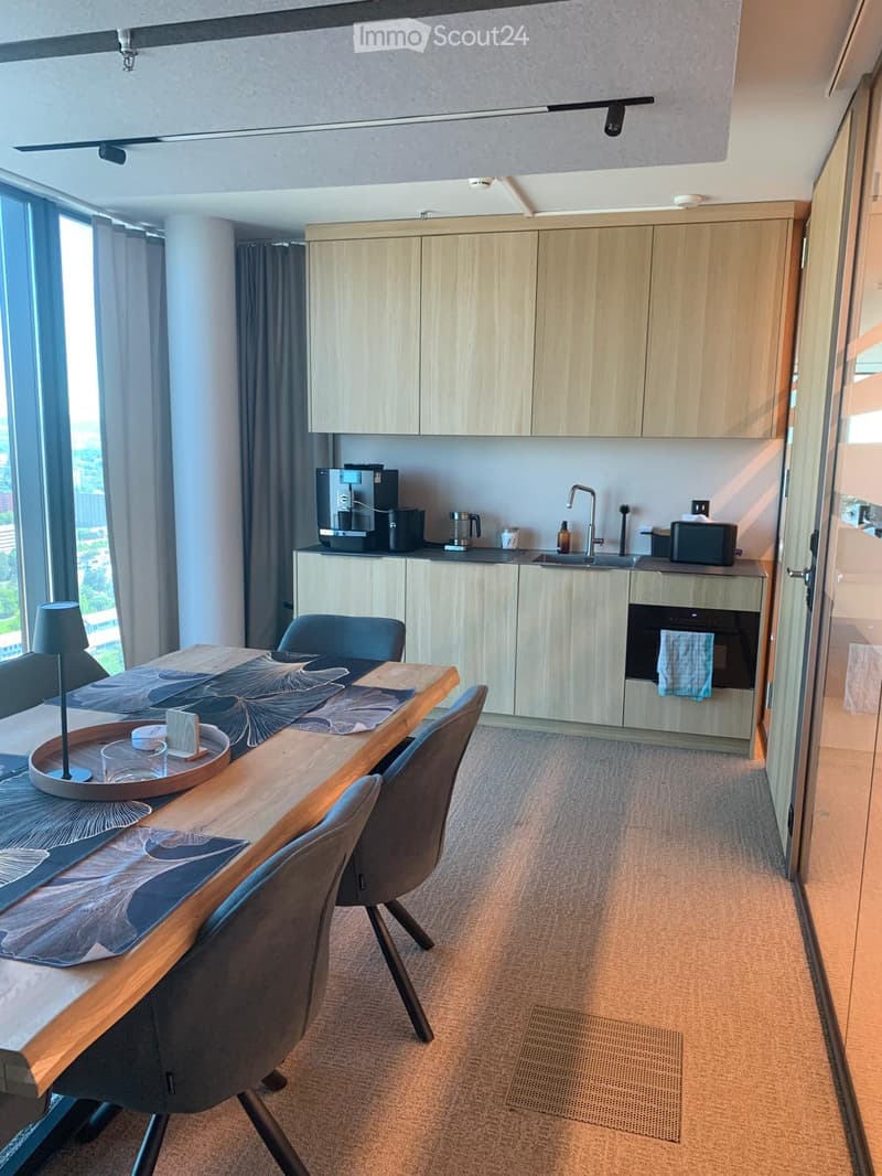Modern kitchen and dining area with light wood cabinets, stainless steel appliances, a long wooden dining table with chairs, and a lamp on the table.