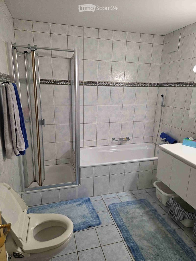 Bathroom with tiled walls and floor, featuring a toilet, bathtub, sink, towels, and bathmats.