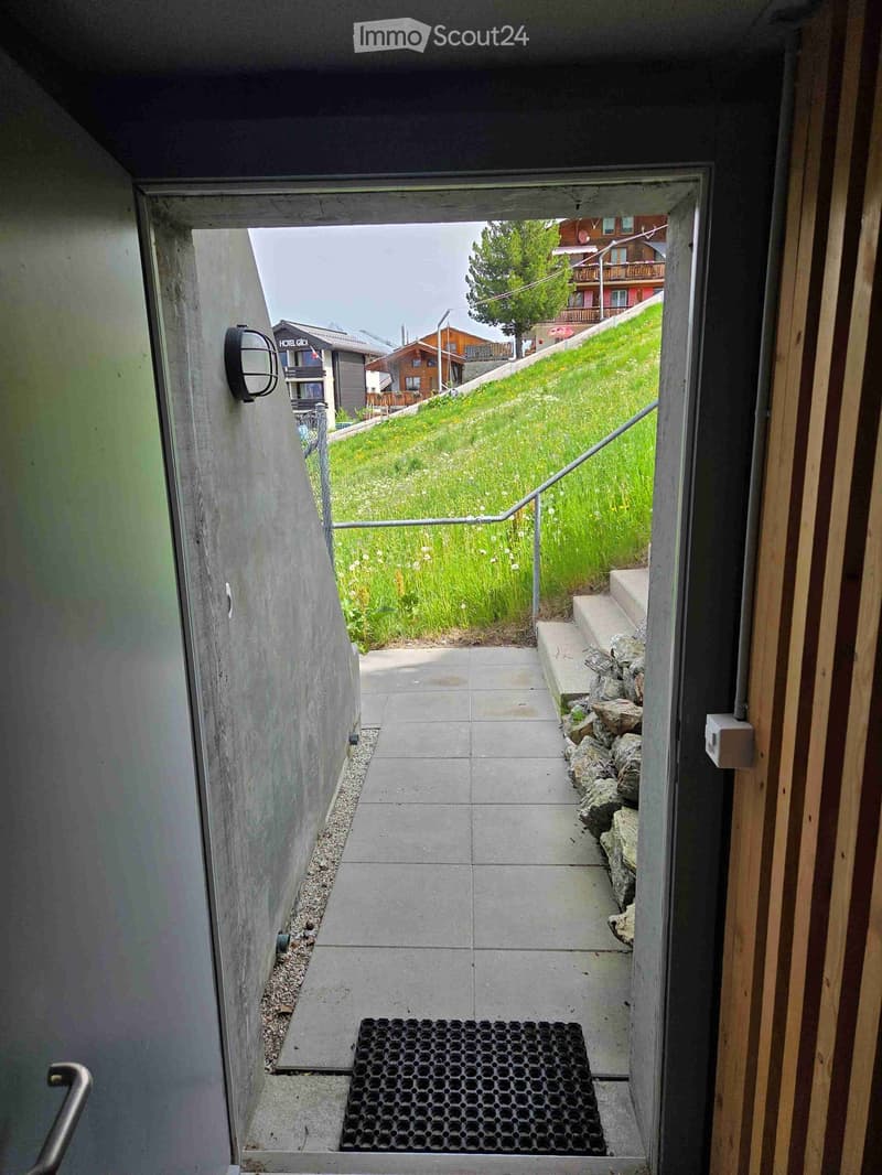 A narrow, tiled hallway leading to an outdoor area with a grassy slope, steps, and a metal railing.