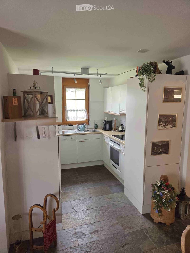 Bright kitchen with white cabinets, a sink, oven, refrigerator, coffee maker, and teapot. Features a window with a curtain, a wooden chair, and a flower pot on the floor.