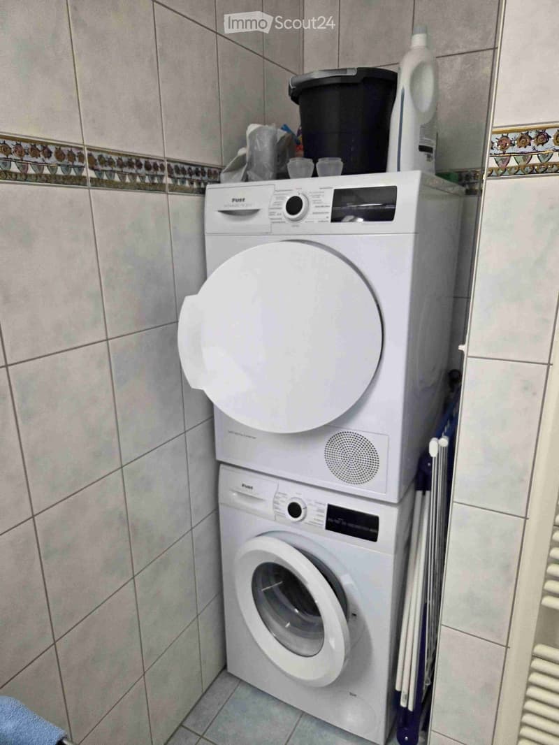 A compact bathroom with a stacked washing machine and dishwasher unit, tiled walls, and cleaning supplies nearby.