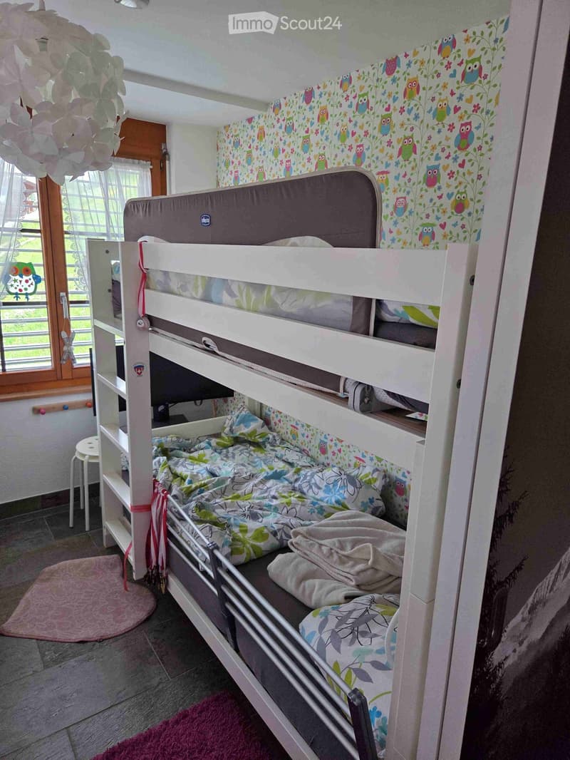 A cozy children's bedroom with a bunk bed, colorful owl-themed wallpaper, a small stool, and a decorative hanging ornament.
