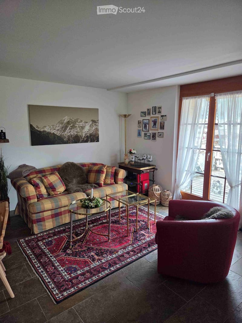 A cozy living room with a plaid-patterned sofa, a red armchair, a small round coffee table, a larger wooden table with a vase and candles, a floor lamp, and several picture frames on the wall. The room features a colorful rug and large windows with curtai