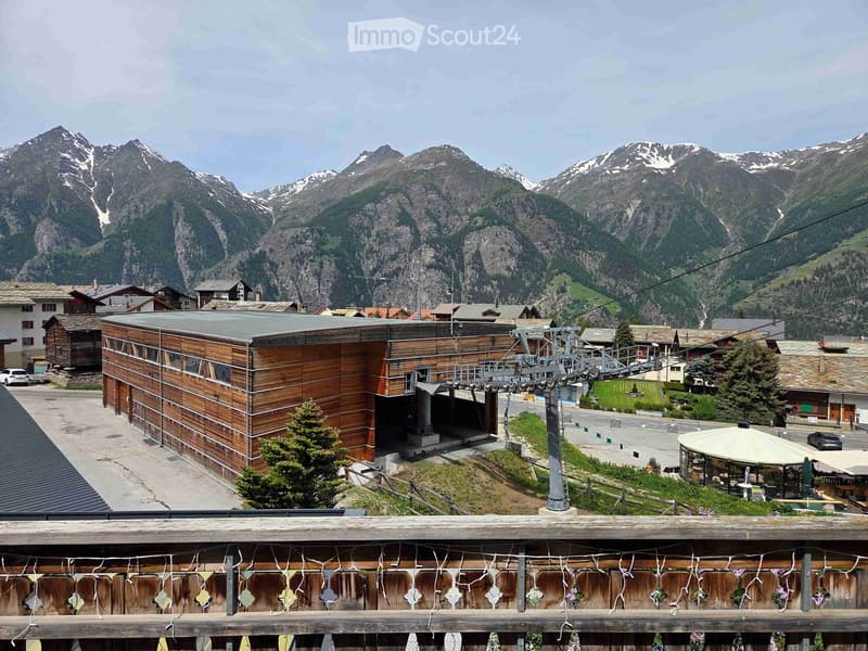 A modern wooden building with a flat roof and large windows, situated in a scenic mountain valley with snow-capped peaks in the background. The building has a cable car station attached to it, and there are several other houses and a small outdoor seating