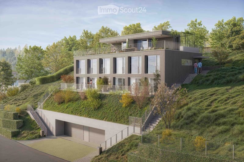 Modern two-story house with a flat roof, large windows, and a balcony on the upper level. The house is situated on a hillside with a landscaped garden and a covered garage at the base.