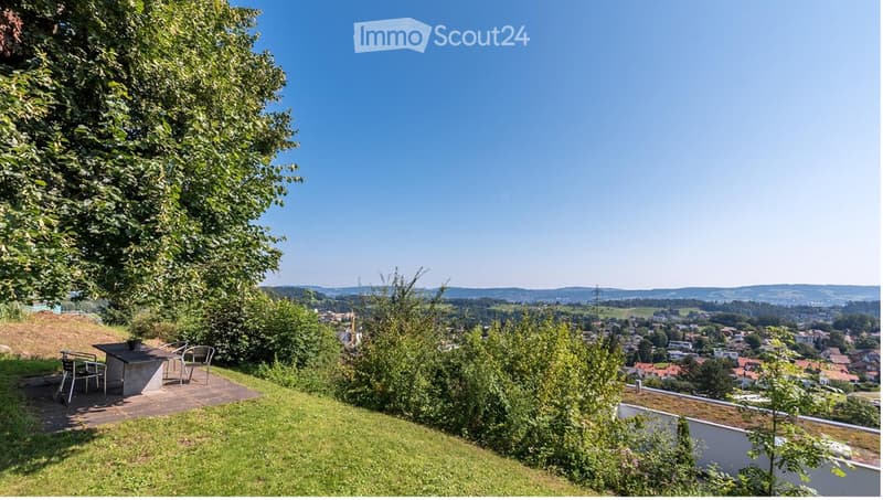 A serene garden or outdoor area with a table and chairs, surrounded by lush greenery and offering a panoramic view of a town and distant hills.