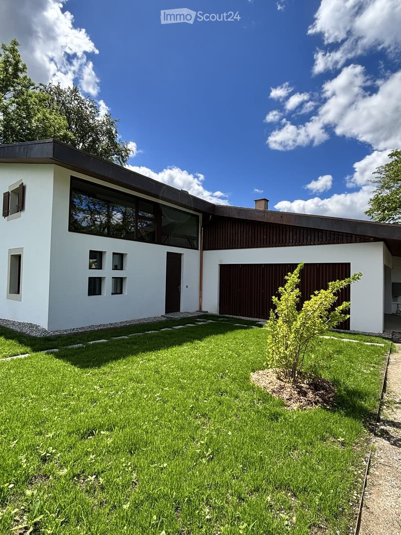 Modern single-story house with white walls, large windows, and a flat roof. Features a garage and a small garden with a tree and stepping stones.