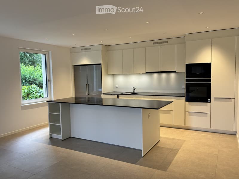 Modern kitchen with white cabinets, black countertops, a central island, and stainless steel appliances including a refrigerator, microwave, and oven. Large window provides natural light.