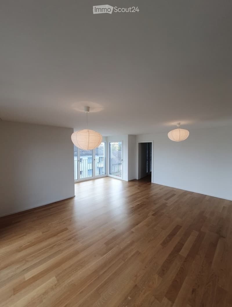 Empty room with wooden flooring, white walls, and two hanging lamps. There is a doorway leading to another room and a large window with a view of the outside.