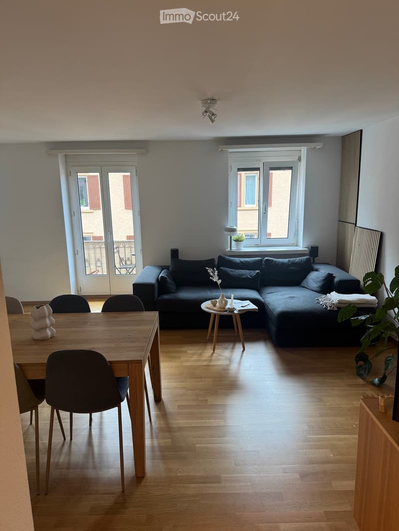 Modern living and dining area with a dark grey sectional sofa, wooden dining table with chairs, small coffee table, and decorative items.