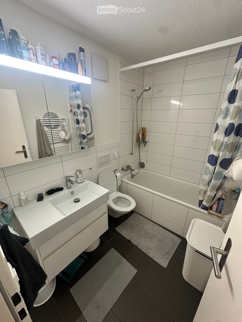 Modern bathroom with white tiles, a sink, toilet, bathtub, and shower area. Includes a mirror with built-in lighting, a towel rack, and a shower curtain with a blue and white pattern.