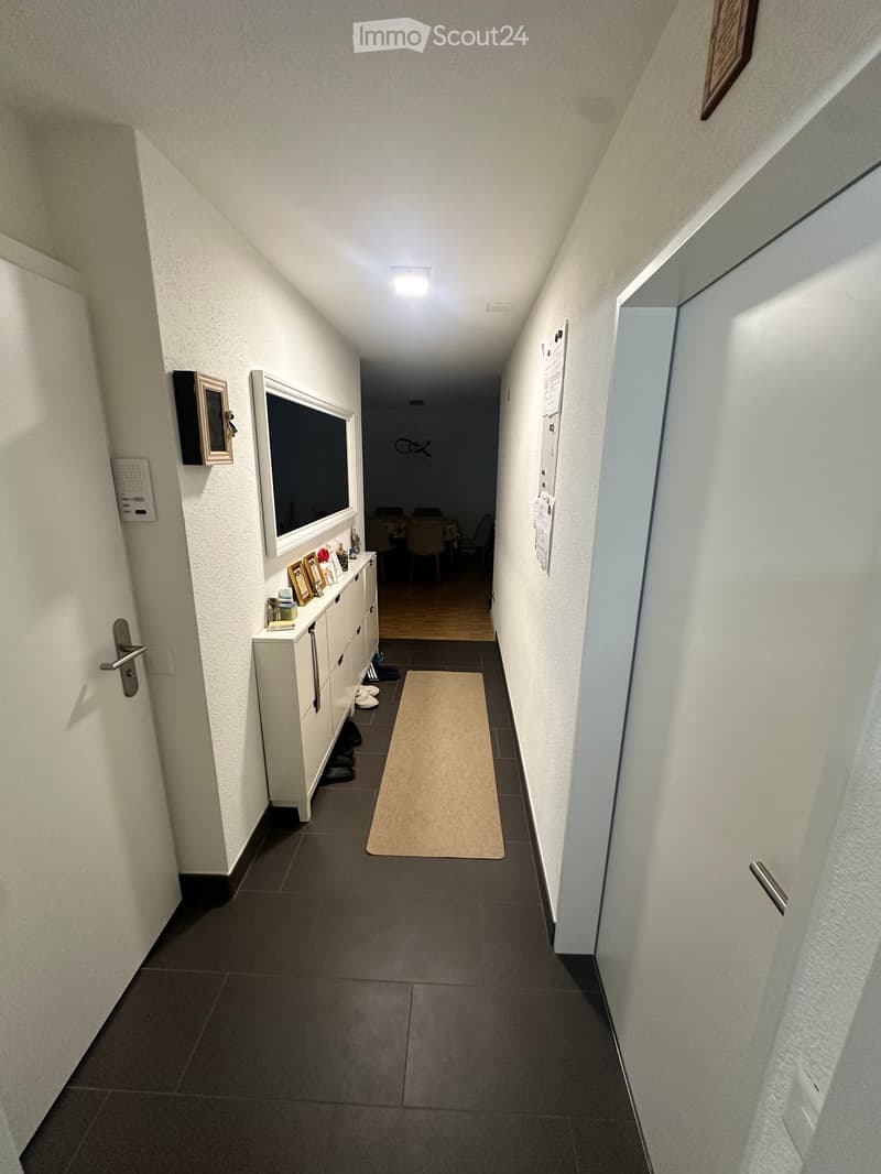 A narrow hallway with white walls, dark tiled floor, and a small rug. A white cabinet with drawers and framed pictures is on the left, and a partially visible room with chairs and a table is at the end.