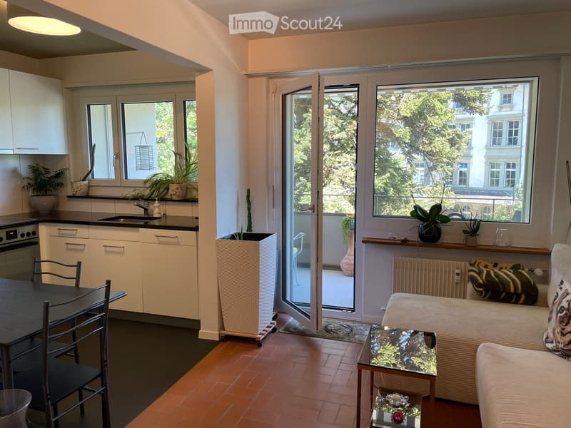 Bright, open-plan living and dining area with a sofa, coffee table, dining table, chairs, kitchen area with oven and sink, and large windows opening to a balcony.