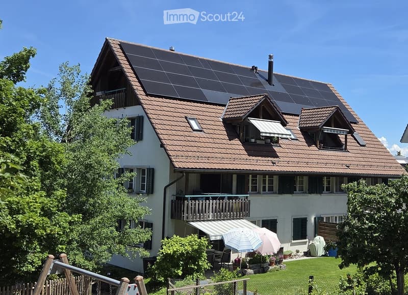 Two-story house with a brown tiled roof, multiple windows, balconies, and solar panels on the roof. The house is surrounded by a well-maintained garden with outdoor furniture, umbrellas, and greenery.