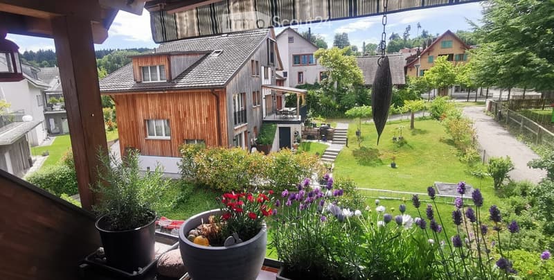 Balcony overlooking a well-maintained garden with various potted plants and flowers, with neighboring houses and greenery visible in the background.