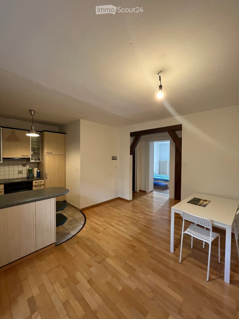 A modern, open-plan living and dining area with wooden flooring, white walls, and a small kitchen area featuring cabinets and a countertop. A white dining table with chairs is positioned near the entrance to a bedroom.