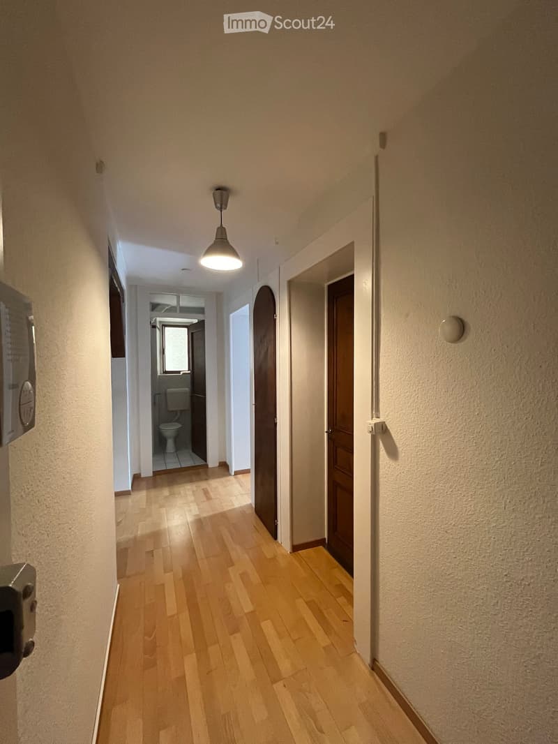 A narrow hallway with wooden flooring, beige walls, and a single hanging light fixture. A partially open door leads to a bathroom with a toilet visible.