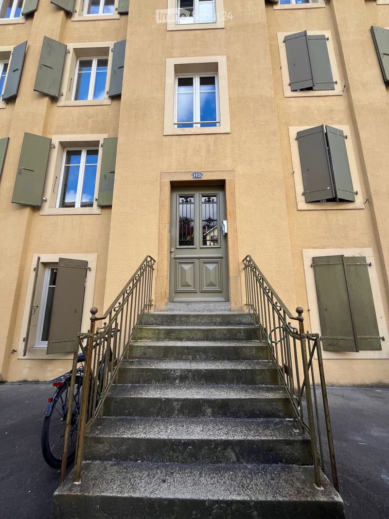Facade of a multi-story building with beige walls, multiple windows with shutters, and a central entrance door with decorative glass panels. A staircase leads up to the entrance.