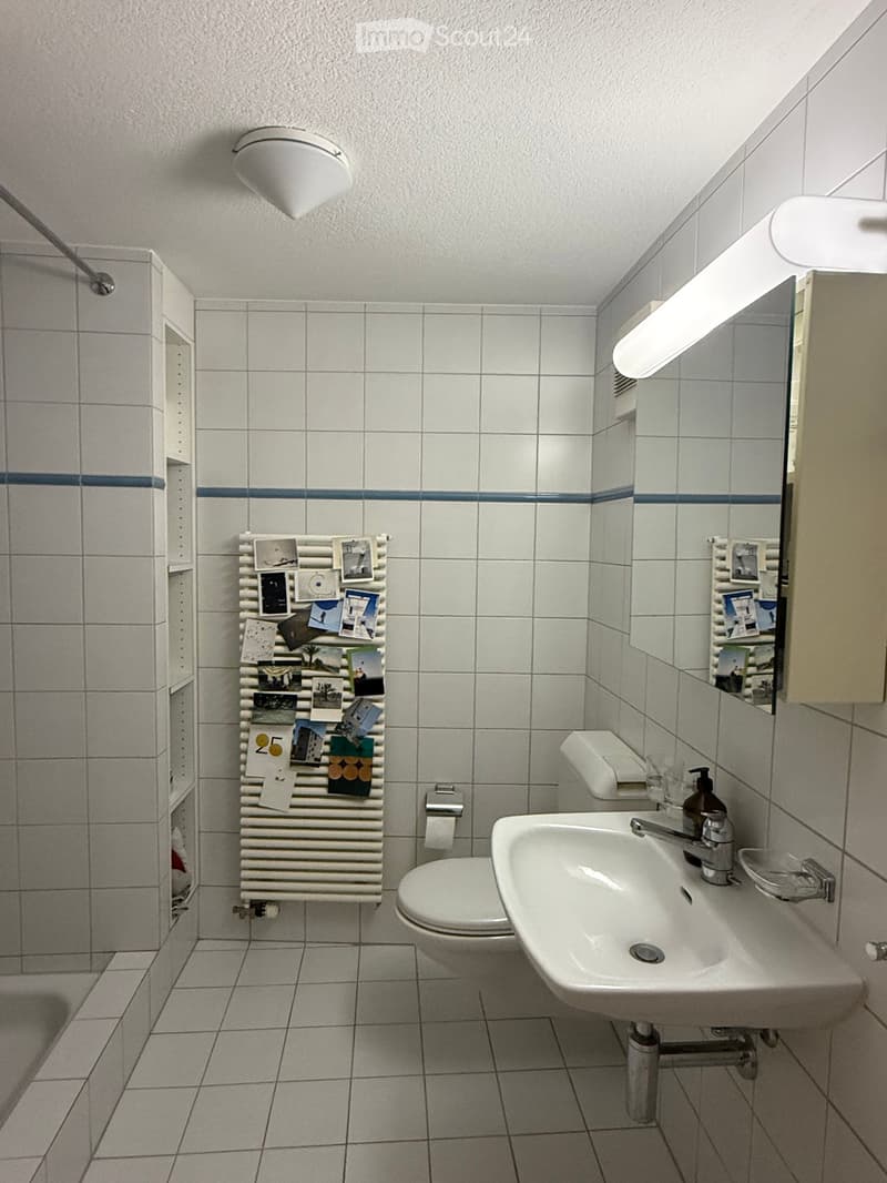 Bathroom with white tiles, a toilet, a sink with a mirror, a radiator with photos, and a shower area with a glass door.