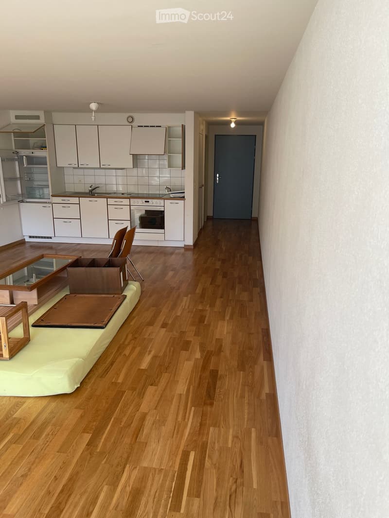 A modern, open-plan living and dining area with wooden flooring, a sofa, a dining table, and a kitchen with white cabinets and stainless steel appliances.