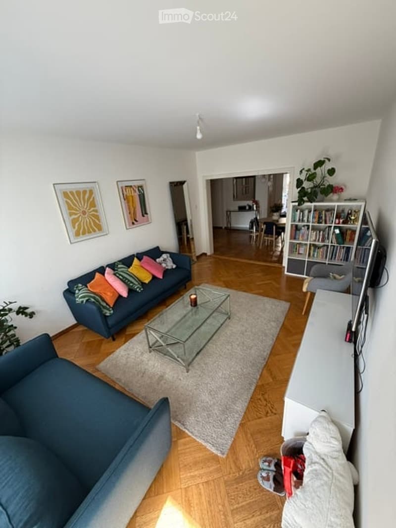 Bright living room with wooden flooring, blue sofa with colorful pillows, glass coffee table, bookshelf, and artwork on the walls.