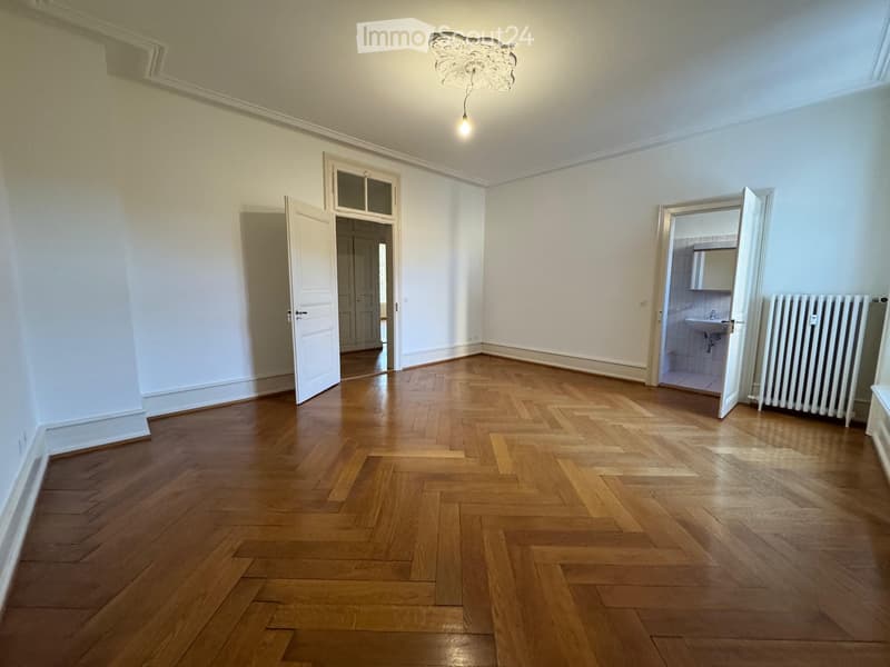 Empty room with wooden herringbone flooring, white walls, and a decorative ceiling light. Two open doors lead to adjacent spaces, one showing a bathroom with a sink and radiator.