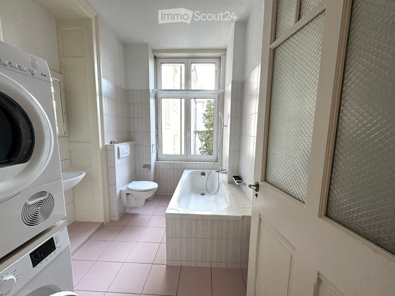 Bathroom with a bathtub, toilet, sink, and washing machine. The room has tiled walls and floor, with a window providing natural light.