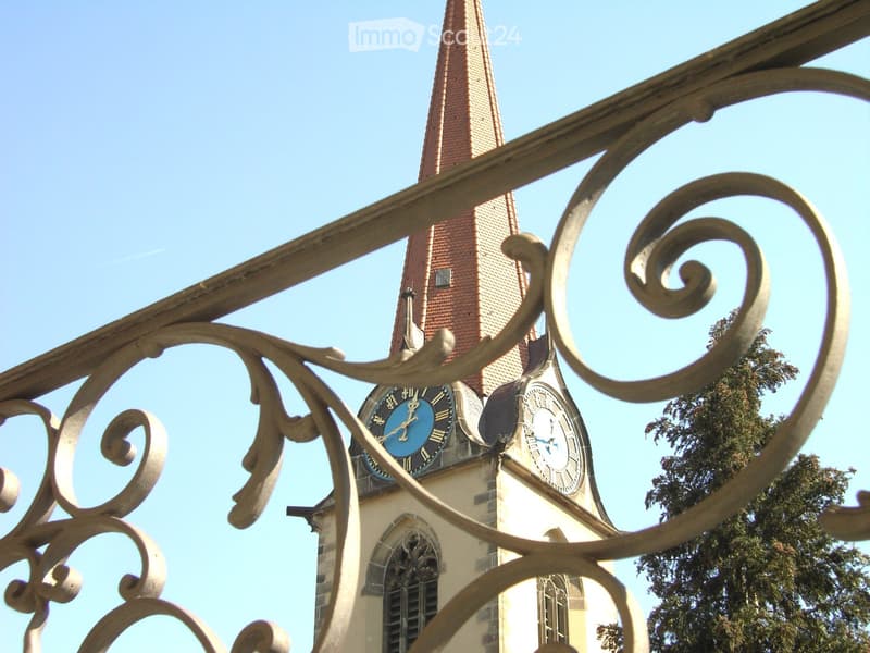 A view of a church tower with a clock, seen through ornate metal railings. The tower has a pointed roof and is partially obscured by the railing.