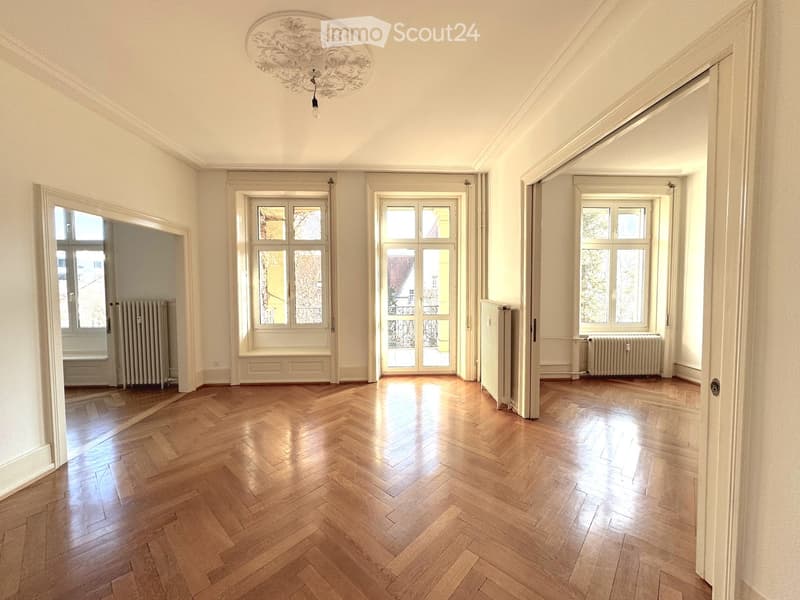 Empty room with wooden herringbone flooring, white walls, large windows, radiators, and an ornate ceiling light fixture.