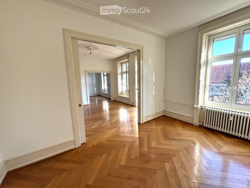 Empty room with wooden herringbone flooring, white walls, large windows, and a radiator. The room has an open doorway leading to another room with similar flooring and white walls.