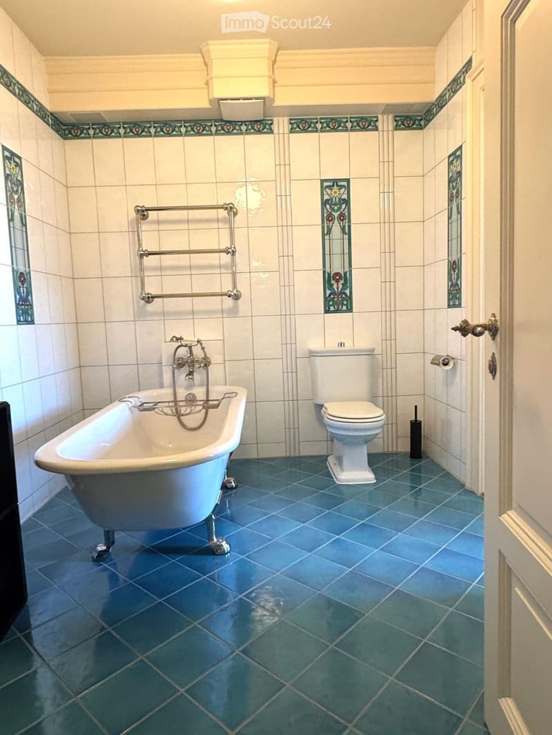 Bathroom with a blue tiled floor, white tiled walls, a bathtub, a toilet, a towel rack, and a toilet paper holder. The walls feature decorative tiles and stained glass panels.
