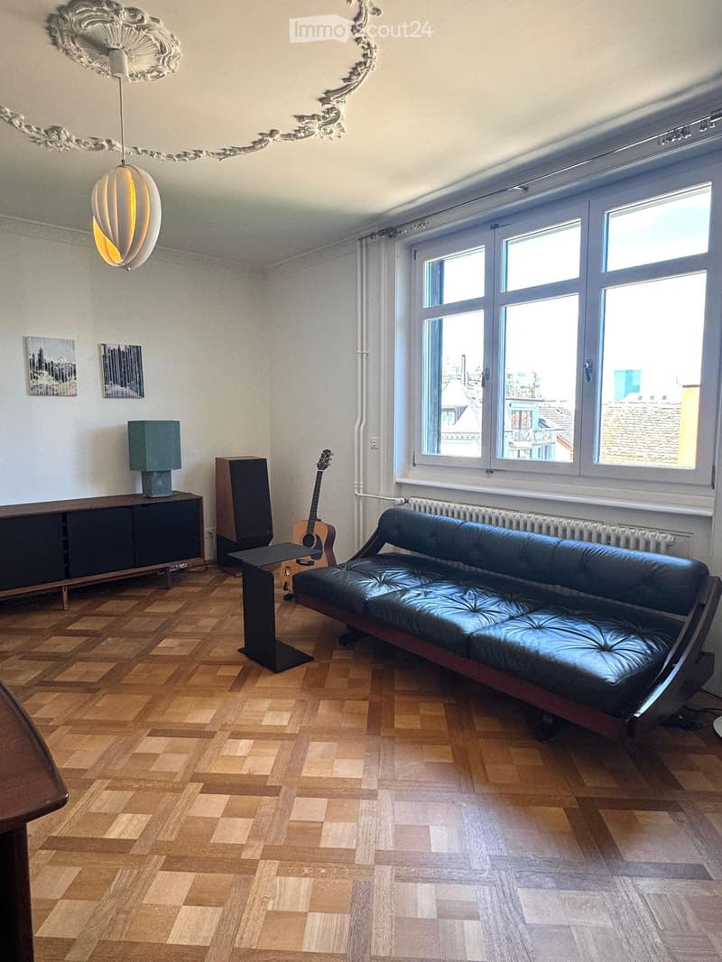 Modern living room with wooden flooring, large windows, a black leather sofa, a guitar, a small table, a lamp, a cabinet, and a speaker.