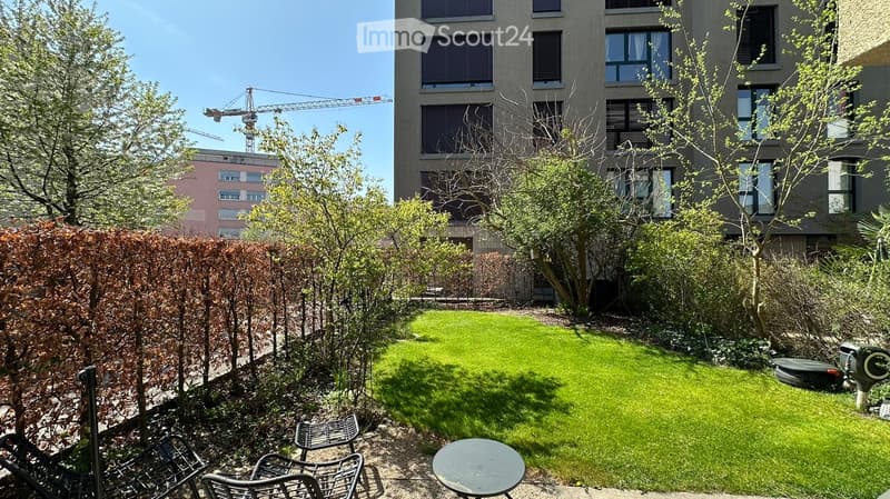 small garden with green grass, trees, shrubs, chairs, table, and a building in the background