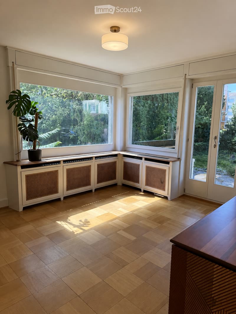 Bright, empty room with wooden flooring, large windows, built-in radiator covers, and a modern ceiling light fixture.