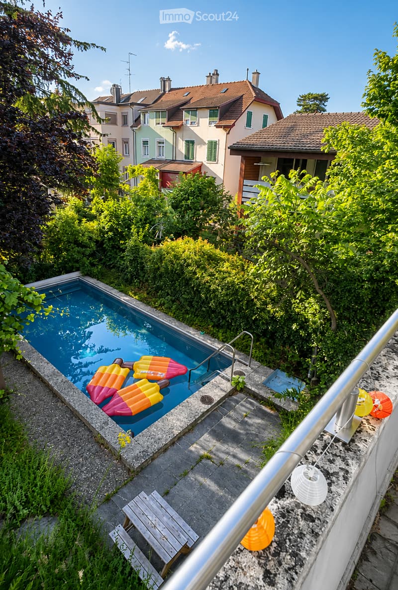 A small swimming pool surrounded by greenery, with inflatable ice cream-shaped floats in the water, a wooden bench, and decorative lanterns nearby.
