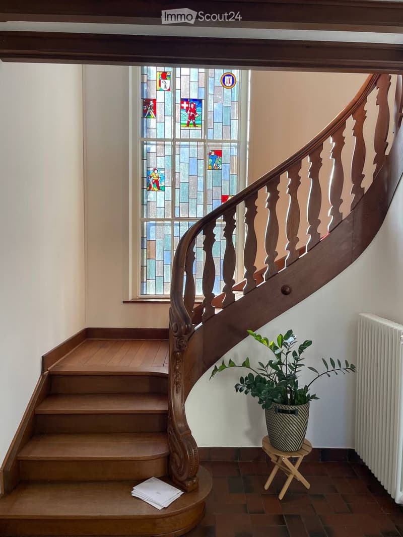 Spiral staircase with wooden handrail and stained glass window, small plant on a stool, and papers on the stairs.