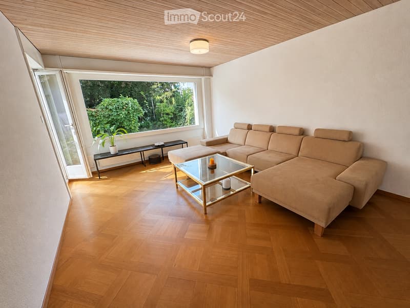 Modern living room with light wooden flooring, a large window with a view of greenery, a beige sectional sofa, a glass coffee table, a side table with a plant, and a small candle on the coffee table.