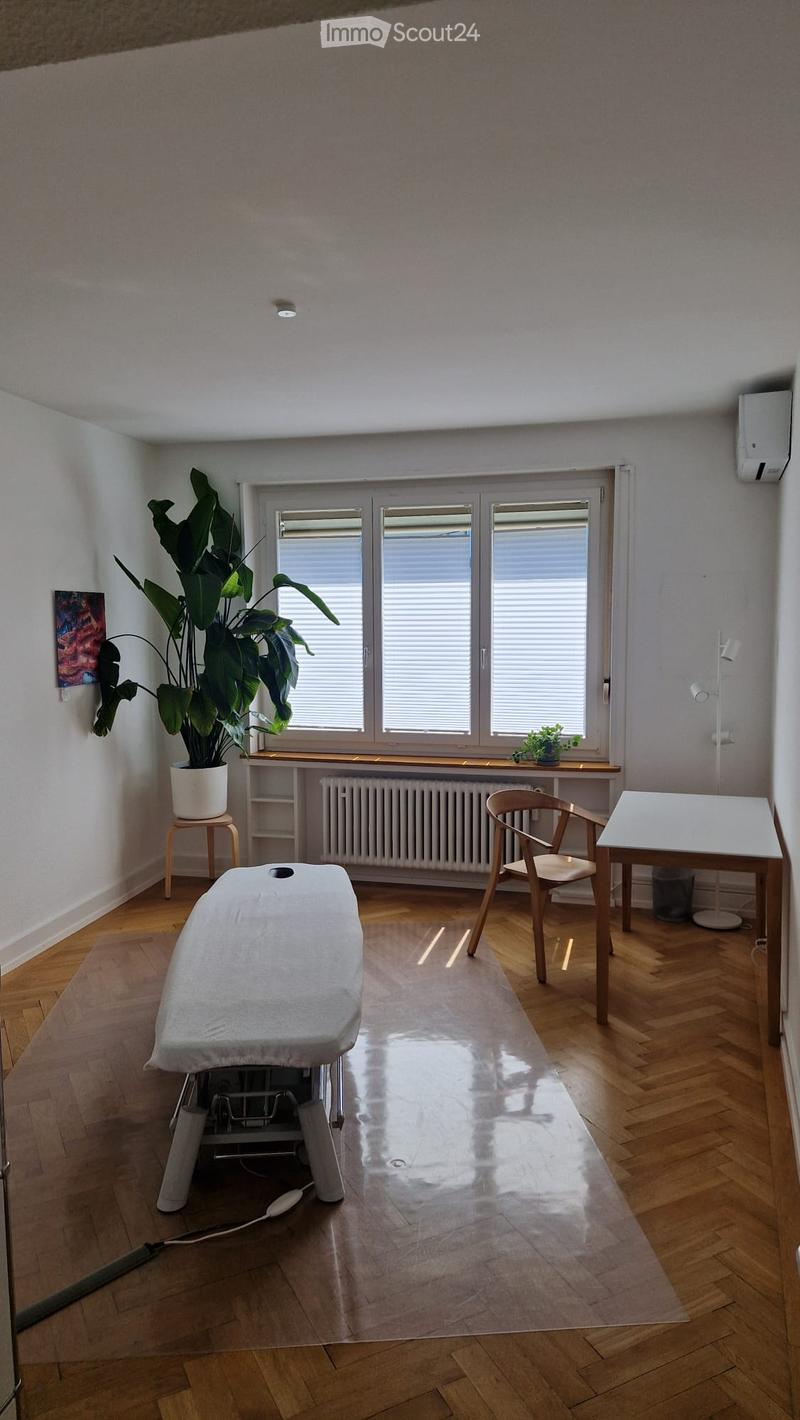Bright room with wooden flooring, large window with blinds, a massage table, a chair, a small table, a plant on a stool, and a radiator under the window.