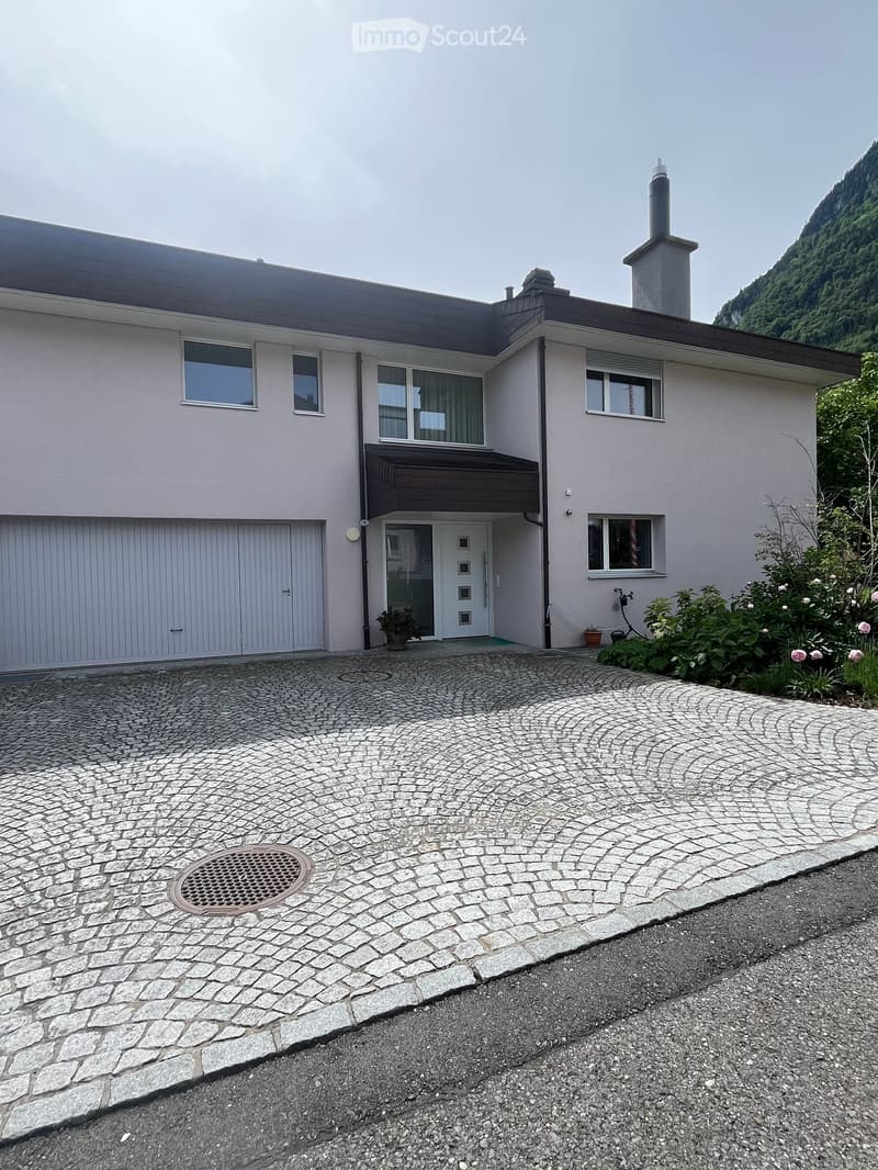 Two-story house with light gray exterior, garage, multiple windows, and a stone-paved driveway.