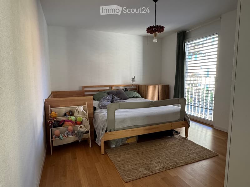 Cozy bedroom with a wooden bed, nightstand, toy basket, and a small rug on the floor. Large window with blinds allows natural light to enter.