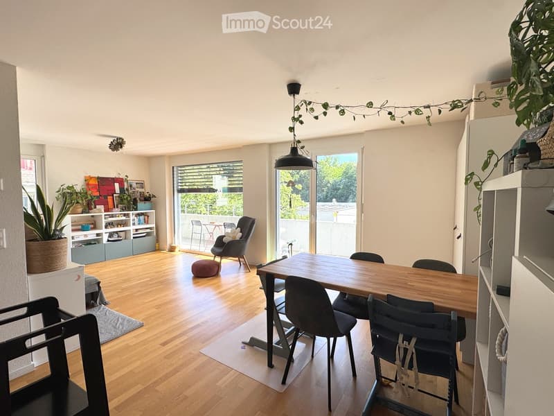 Bright living and dining area with wooden flooring, large windows, and a balcony. Includes a dining table with chairs, a sofa, a shelf with plants and books, and a hanging pendant light.