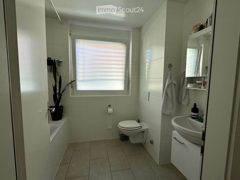 Modern bathroom with tiled walls and floor, featuring a toilet, sink, bathtub, and a small window with blinds. A towel hangs on a rack, and a potted plant sits on the edge of the bathtub.
