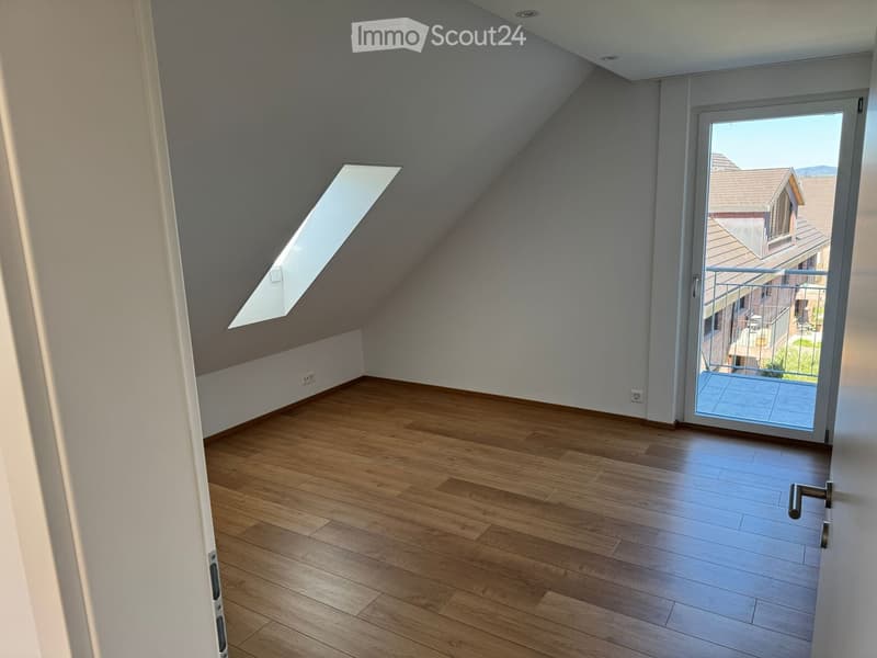 Empty attic room with wooden flooring, white walls, skylight window, and sliding glass door leading to a balcony.